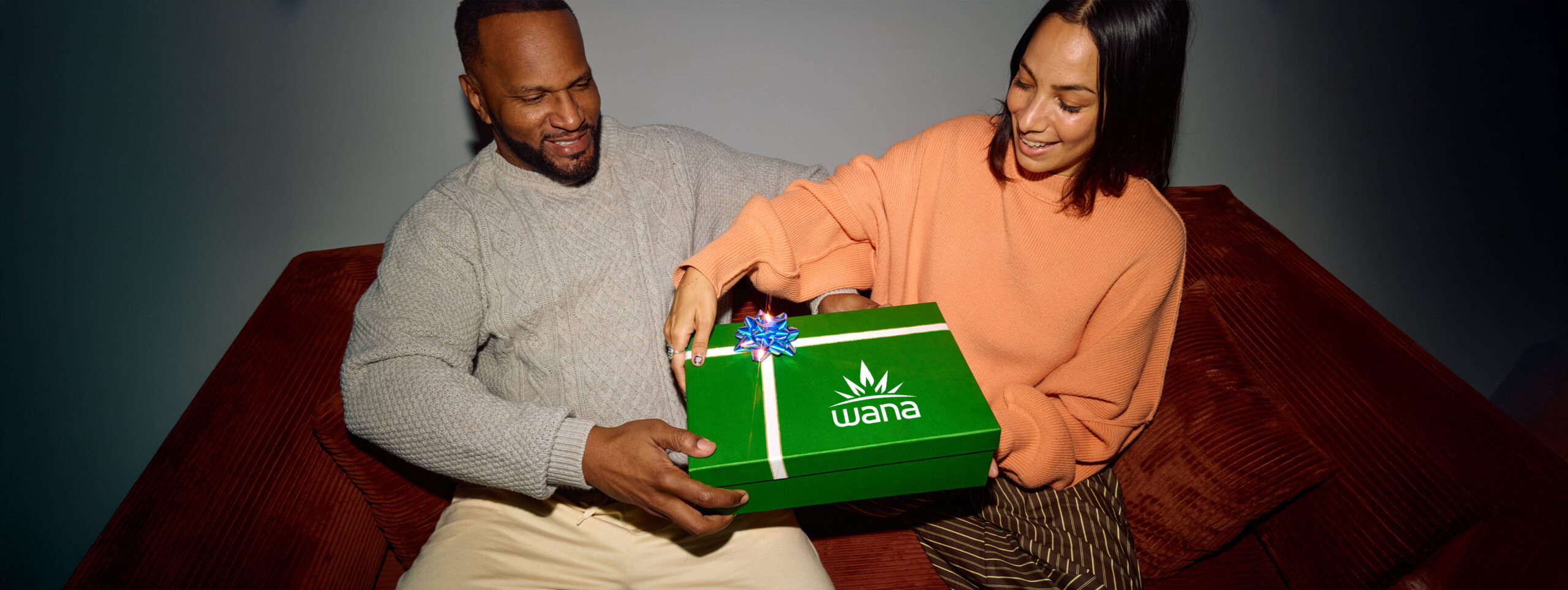 A man handing a gift-wrapped green box marked with the Wana logo to the woman beside him.